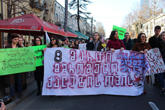 La photo montre une manifestation où une foule de personnes tient une bannière en géorgien qui se lit comme suit : « Le 8 mars pour les femmes travailleuses ».